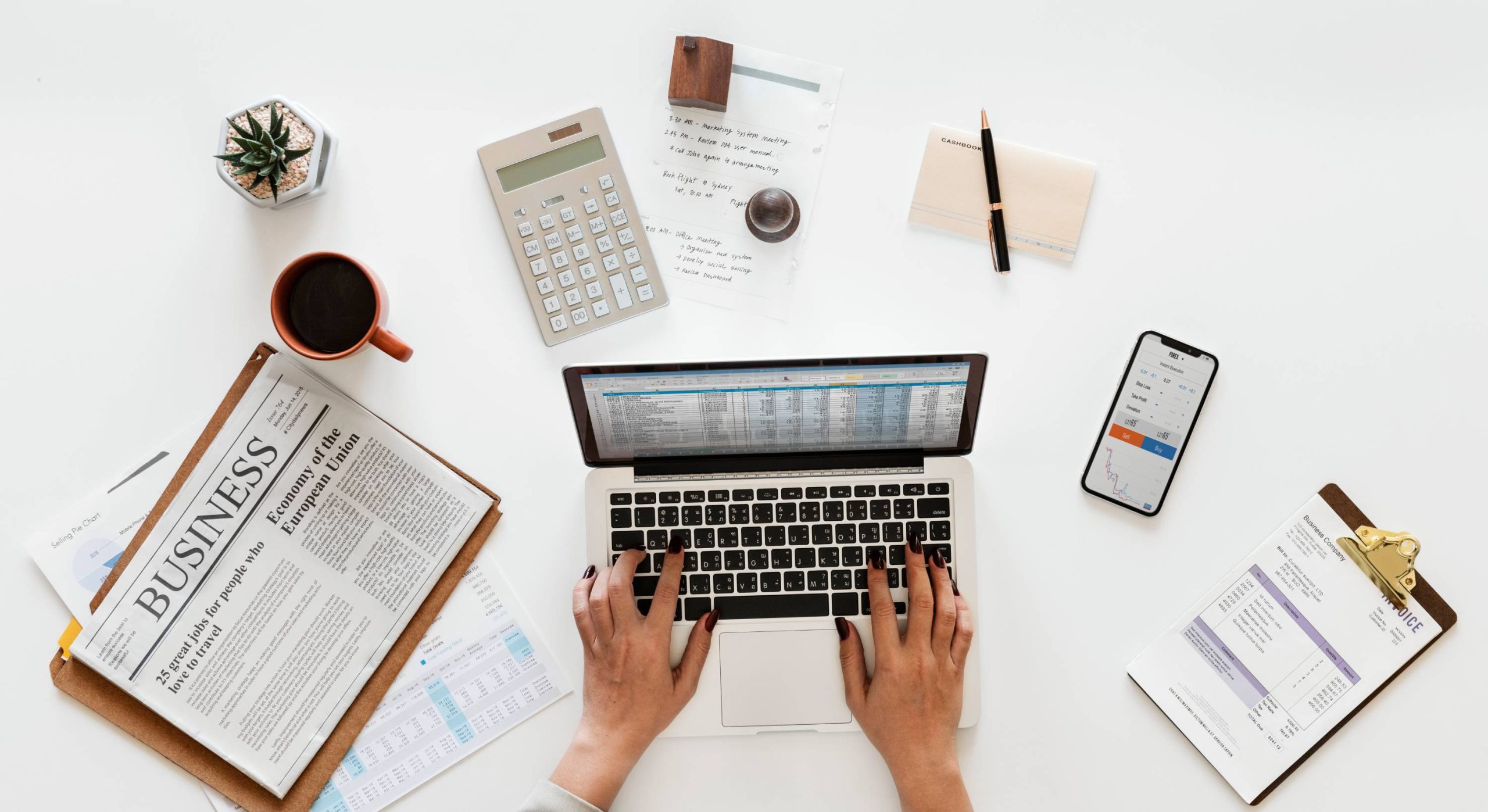 Overhead view of hands typing on a laptop showing a spreadsheet, with a calculator, invoice, and notes on a white desk